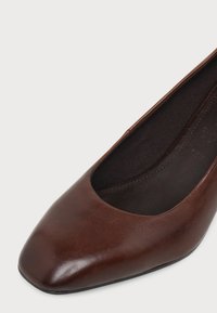Gros plan d'une chaussure plate pour femme en cuir marron avec un bout carré et une doublure intérieure noire sur fond blanc.