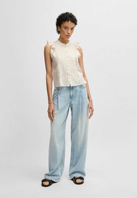 Ljust beige ärmlös topp med volangdetaljer och vertikala veck, kombinerad med ljusblå vida jeans och svarta sandaler.