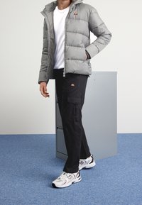 Grijze pufferjack met capuchon, witte t-shirt, zwarte cargo-broeken en witte sneakers met rode accenten, tegen een lichte achtergrond.