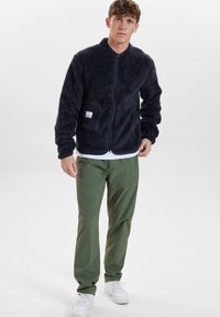 Giacca in pile blu navy con zip frontale, colletto alto e taschino sul petto; abbinata a pantaloni verde oliva e sneakers bianche.