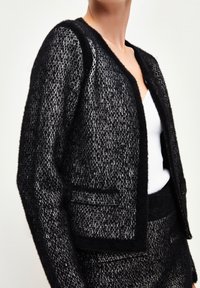 Svart och vit texturerad kavaj med rundad halsringning, utan knappar, och två framfickor, med en mjuk, fluffig yta och kortad design.