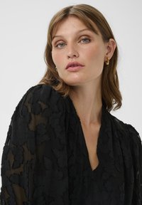 Schwarzes transparentes Kleid mit floralem Muster, tiefem V-Ausschnitt und puffartigen Ärmeln. Modell trägt goldene Creolen, dezentes Make-up und einen neutralen Hintergrund.
