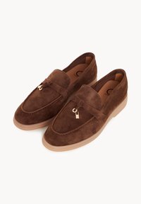 Paire de mocassins en daim marron avec semelles beige clair et petits glands décoratifs sur le dessus, présentés sur un fond blanc.