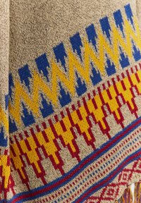 Textil țesut cu o bază bej, având modele intricate în nuanțe de albastru, galben și roșu, prezentând motive geometrice și accesorii cu franjuri.