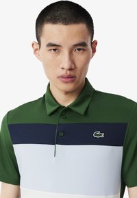 Grön polotröja med horisontella ränder i marinblått, ljusblått och vitt. Tre-knapps knäppning och broderad logotyp på bröstet.