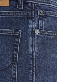 Tamsiai mėlynos džinsinės kelnės turi klasikinį penkių kišenių dizainą, matomus siūles, metalinius mygtukų akcentus ir tekstūruotą apdorojimą.