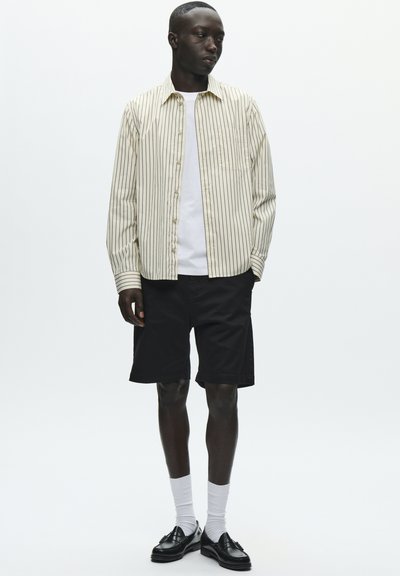 Chemise à boutons rayée en crème et noir, associée à un short noir. Chaussettes blanches montantes et chaussures noires complètent le look. Design simple et moderne.