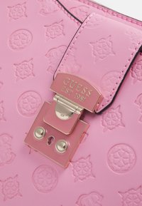 Vaaleanpunainen teksturoitu käsilaukku, jossa on kohokuvioitu logokuvio, metallinen lukko ja kullanväriset yksityiskohdat.