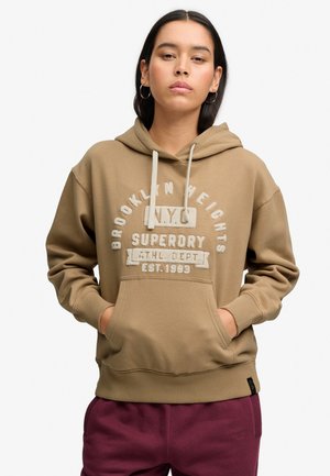 Kobieta ubrana w beżową bluzę z kapturem z napisem "Brooklyn Heights N.Y.C. Superdry Athl. Dept Est. 1983" i bordowe spodnie, stojąca z rękami w kieszeniach.