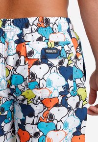 Bañador colorido con un patrón de Snoopy en azul, naranja y verde sobre un fondo blanco, con un bolsillo trasero y una etiqueta de PEANUTS.