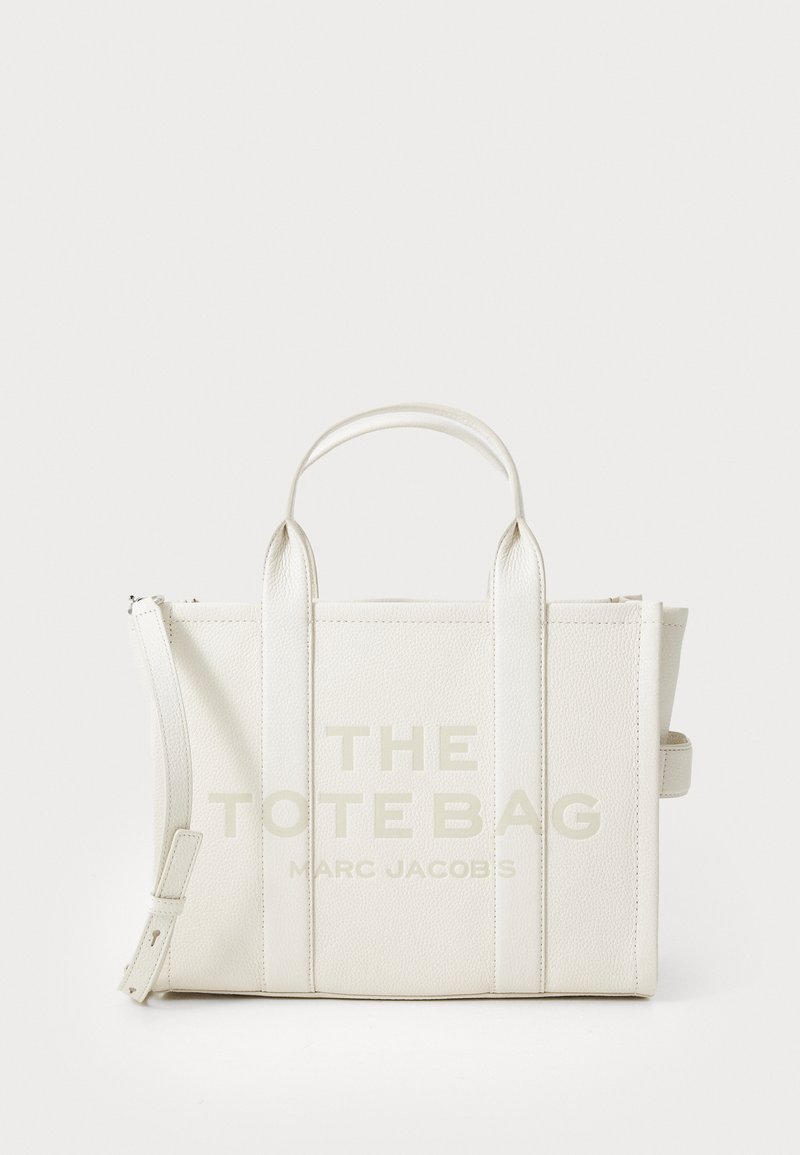 Saco tote de couro branco texturizado com duas alças e fecho com zíper. Apresenta um grande texto impresso "THE TOTE BAG" e "MARC JACOBS" na parte da frente.