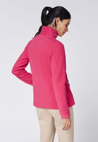 Woman with black ponytail wearing pink fleece jacket and beige pants, standing with her back slightly turned to the camera.