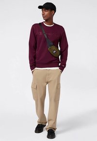 Sudadera granate con logo bordado, pantalones cargo beige con bolsillos laterales, zapatos negros y bolso cruzado verde. Estilo casual y cómodo.