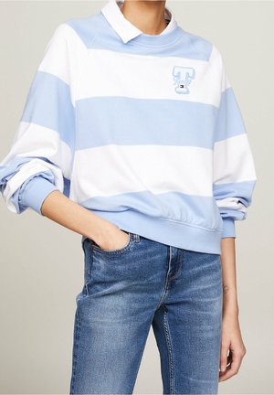 Persoon draagt een blauw-wit gestreepte trui met brede strepen en een klein logo op de borst, gecombineerd met een casual blauwe spijkerbroek, één hand in de zak.