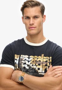 Camiseta negra con cuello crema que presenta un gráfico dorado y blanco desgastado. Se lleva con un reloj dorado y una pulsera de cuero en la muñeca.