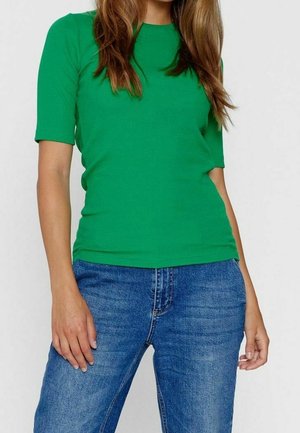 Femme portant un t-shirt vert ajusté à manches courtes, rentré dans un jean bleu, debout devant un fond blanc uni.