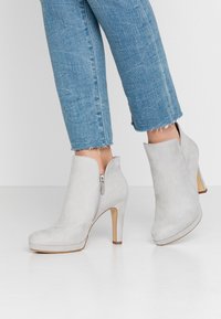 Graue Wildleder-Stiefeletten mit einem seitlichen Reißverschluss, hohem schlanken Absatz, runder Zehenpartie und glatter Textur. Kombiniert mit knöchellangen blauen Jeans.