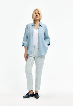 Blonde Frau steht mit der Hand in der Tasche, trägt ein hellblaues Hemd über einem weißen Top, gestreifte Hosen und dunkelblaue Loafers.