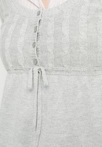 Ανοιχτό γκρι πλεκτό ένδυμα με σχέδιο καλω�δίου, πέντε κουμπιά στο κέντρο και κορδόνι που δένεται κάτω από τα κουμπιά.