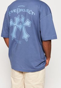 Übergröße blaues Baumwoll-T-Shirt mit einem großen hellblauen Grafikdruck auf dem Rücken, das ein Kreuzdesign undTextdetails in der Nähe des Saums zeigt.