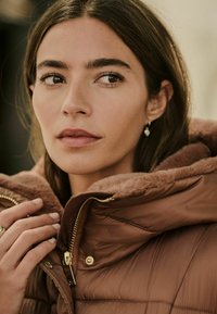 Chaqueta acolchada marrón con cuello de piel sintética, cremalleras doradas y botones a presión. El modelo tiene el cabello largo y está agarrando suavemente el cuello. Diseño minimalista.