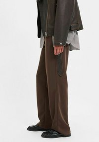 Brūni bikses ar brīvu piegriezumu un sānu kabatām, kombinētas ar tumšu ādas jaku un melnbaltu svītrainu kreklu zem tās.