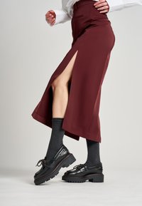 Bordeaux rok met een hoge split, gecombineerd met zwarte leren schoenen met dikke zolen en decoratieve veters, en donkergrijze sokken.