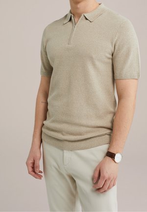 Mann trägt ein beiges kurzärmeliges Strick-Poloshirt mit Reißverschluss, helle Hose und eine braune Lederarmbanduhr am linken Handgelenk.