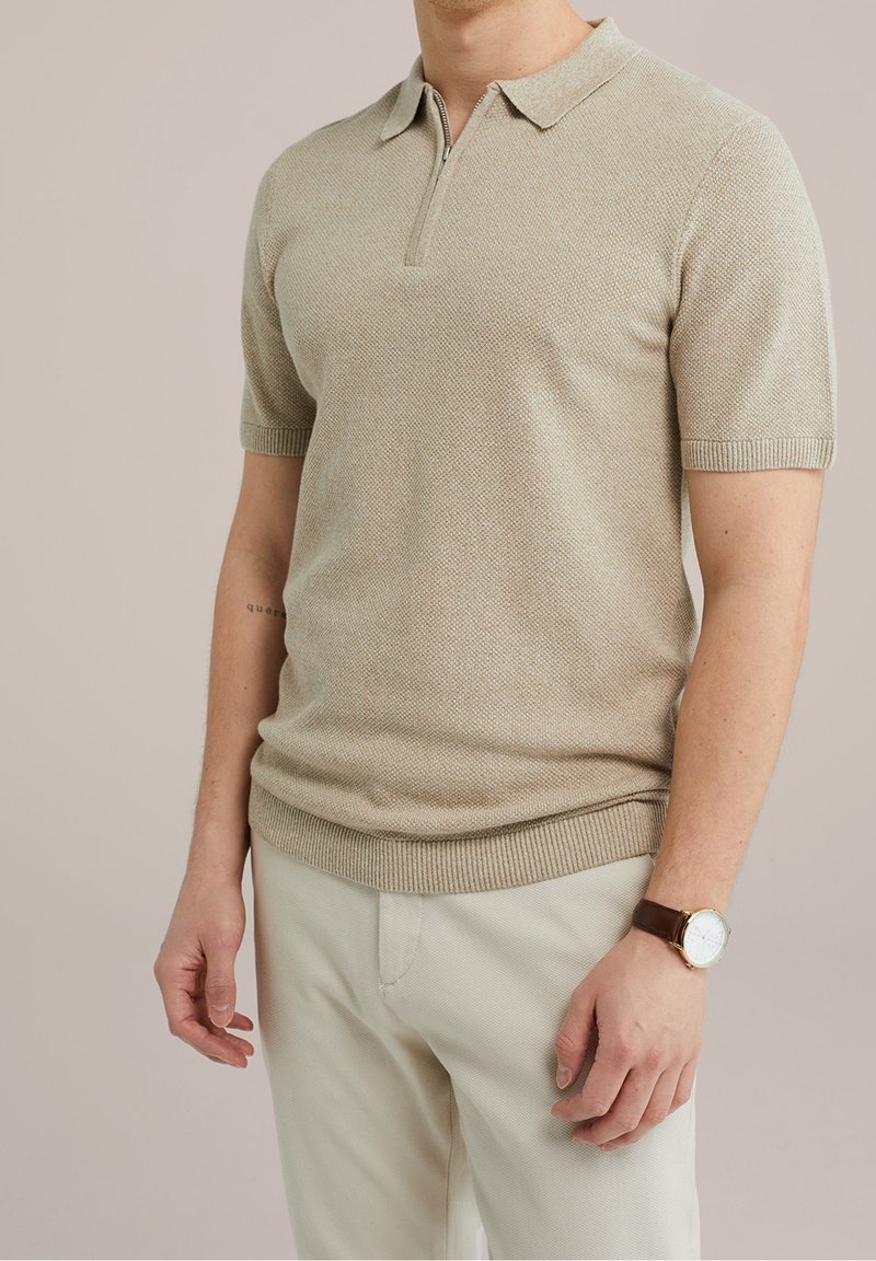 Homme portant un polo en maille beige à manches courtes avec fermeture éclair, pantalon de couleur claire et montre à bracelet en cuir marron au poignet gauche.