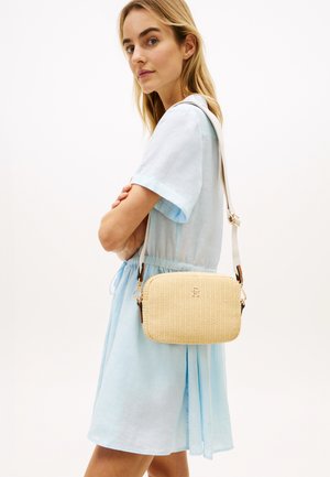 Femeie în rochie albastru deschis care poartă o geantă crossbody bej cu textură și curea reglabilă pe umăr.