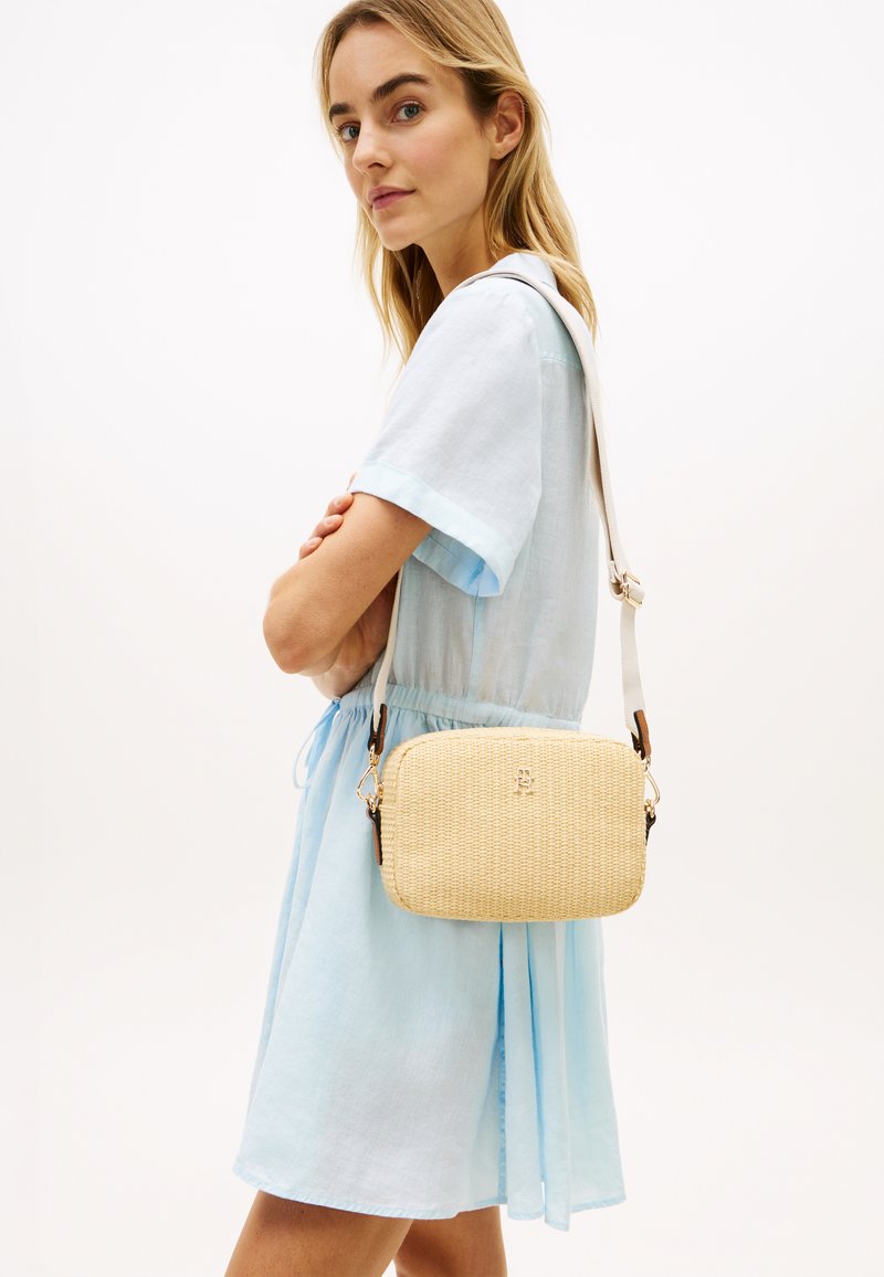 Γυναίκα με ανοιχτό μπλε φόρεμα που κρατάει μπεζ τσάντα χιαστί με υφασμάτινη υφή και ρυθμιζόμενο λουρί στον ώμο.