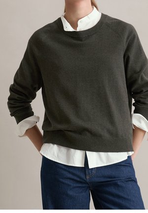 Personne portant un pull gris foncé à col rond sur une chemise blanche boutonnée et un jean bleu, les mains posées sur les hanches.