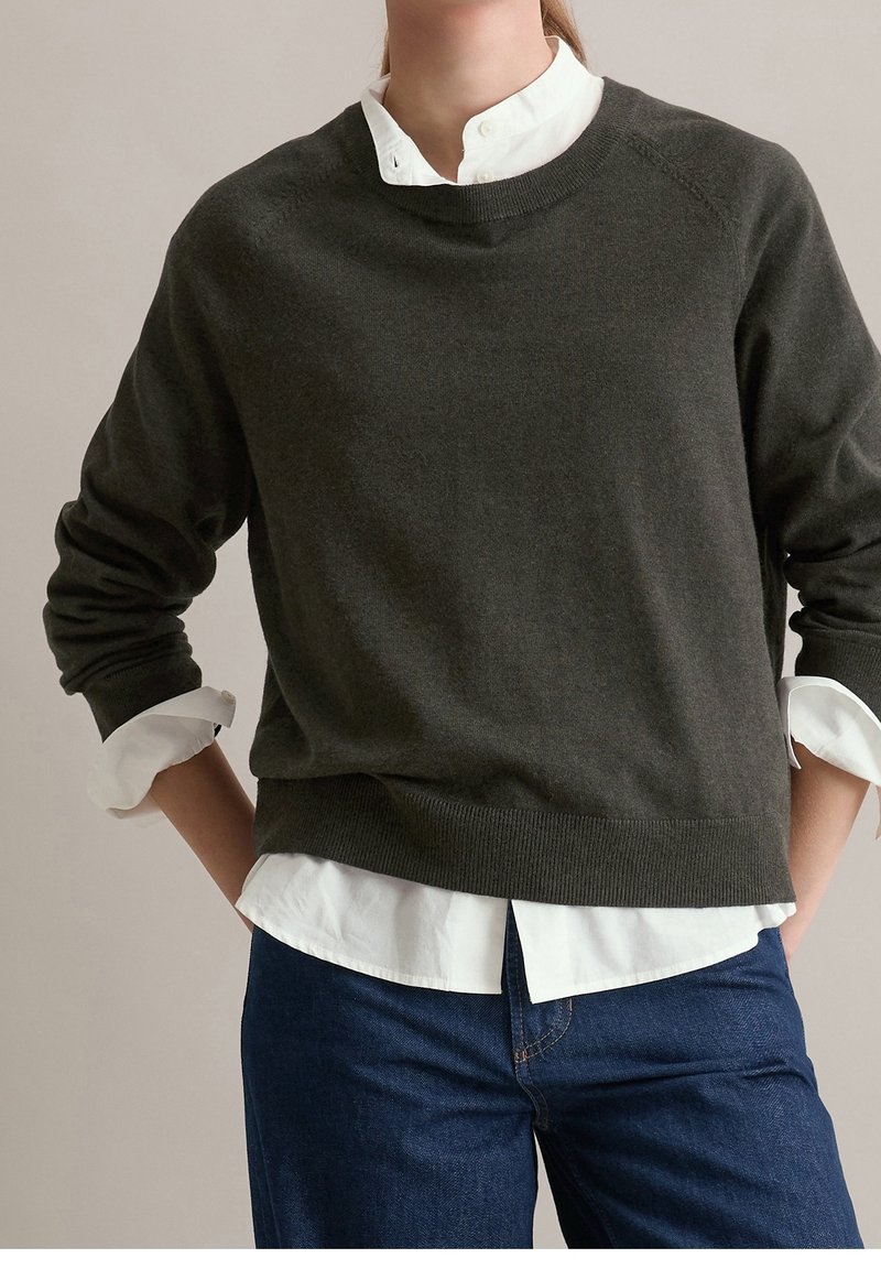 Personne portant un pull gris foncé à col rond sur une chemise blanche boutonnée et un jean bleu, les mains posées sur les hanches.