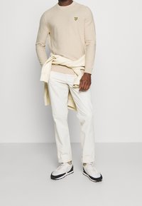 Mann in einem strukturierten beigen Pullover mit Logo, hellen Hosen, Sneakers und einem cremefarbenen Sweatshirt um die Taille vor einfarbigem Hintergrund.