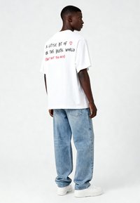 Balts kokvilnas T-krekls ar melnu un sarkanu drukātu ziņu uz muguras. Apvienots ar gaiši ziliem vaļīgiem džinsiem un baltiem sporta apaviem.