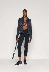 Jaqueta preta desportiva com design texturizado, top de desporto floral, leggings pretos de cintura alta com detalhes florais e sapatos de desporto pretos. Segurando uma raquete de ténis.
