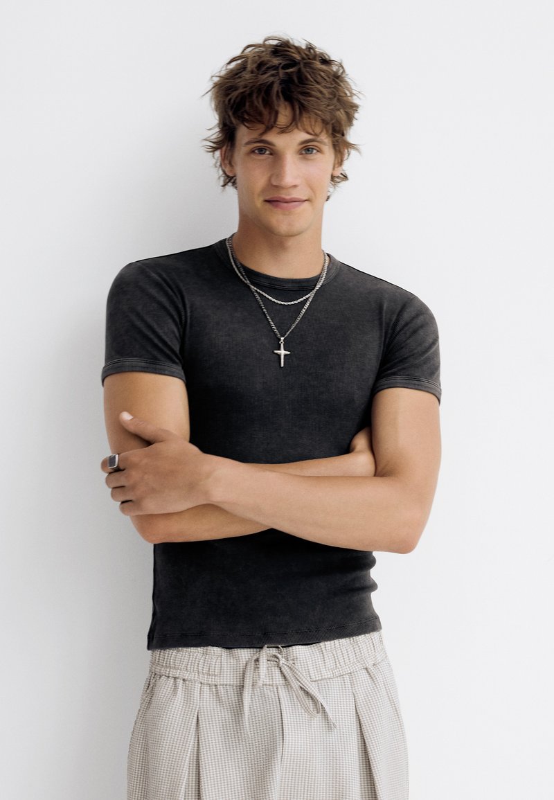 Jeune homme aux cheveux ébouriffés portant un t-shirt noir ajusté, un collier croix en argent et un pantalon large à carreaux clairs, bras croisés.