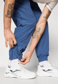 Mėlyni, lengvi, elastiniai kelnių rankogaliai su reguliuojamais apačiais, derinami su baltais sportbačiais. Matomi ryškūs tatuiruotės ant rankų.