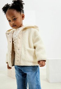 Tejfehér bolyhos fleece dzseki bézs kiemelésekkel, elülső gombokkal és két oldalzsebbel, világoskék farmernadrággal és pöttyös inggel párosítva.