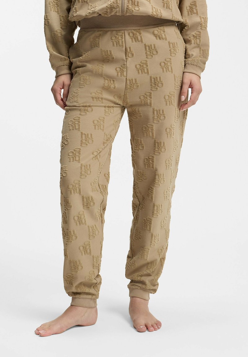 Pantalon de survêtement beige texturé avec des poignets élastiques, présentant un motif de logo en relief répétitif, porté pieds nus par une personne debout.