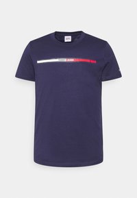 Námorně modré bavlněné tričko s horizontálním grafickým prvkem v bílé a červené barvě, na kterém je uprostřed hrudi nápis "TOMMY JEANS".