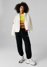 Λευκό φλις μπουφάν με κίτρινο φερμουάρ, μαύροι αθλητικοί παντελόνες με λογότυπο και λευκά αθλητικά παπούτσια με μαύρες λεπτομέρειες, σε ουδέτερο φόντο.