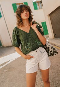Blouse verte brodée avec des manches bouffantes, à pois texturés ; associée à un short en denim blanc avec des coutures bleues. Sac tissé multicolore.