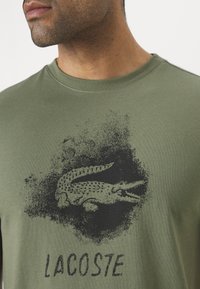 Olive green cotton t-shirt featuring a black, textured crocodile logo and the word "LACOSTE" beneath it. Round neckline and short sleeves.