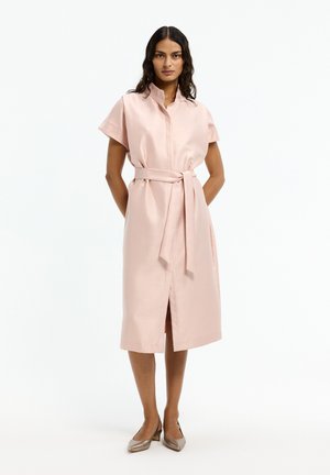 Frau steht mit den Händen hinter dem Rücken und trägt ein hellrosa midikleid mit Gürtel sowie beige Schuhe mit niedrigem Absatz vor weißem Hintergrund.