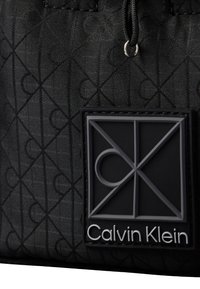 Μαύρο ύφασμα με επαναλαμβανόμενο γεωμετρικό μοτίβο λογότυπου και μια μαύρη και γκρι ετικέτα λογοτύπου Calvin Klein ραμμένη κοντά σε κορδόνι με μεταλλικό οφθαλμό.