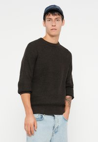 Bruine gebreide trui met een geribbelde structuur, ronde halslijn en driekwartmouwen, gecombineerd met lichtblauwe denim jeans.