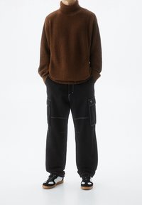 Hnědý žebrovaný svetr s rolákem ve spojení s volnými černými kalhotami typu cargo s bílým prošitím a bočními kapsami, doplněno černobílými teniskami.