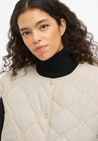 Beežikirju vest, millel on teemantmustriline tikand ja suruaugukinnitused, kantud musta kõrge kaelusega kampsuni peal. Pehme kangas kergelt läikiva viimistlusega.