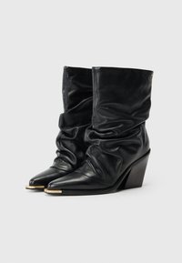 Bottines en cuir noir avec un design fluide, un bout pointu et un talon en bois moyen. Présente des accents en métal doré.